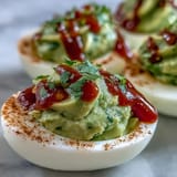 Creamy avocado deviled eggs with a spicy Sriracha drizzle, garnished with fresh cilantro for a zesty party appetizer.