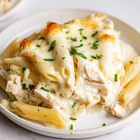 A close-up of the creamy Chicken Ranch Pasta Bake reveals tender penne and shredded chicken coated in a rich, herb-flecked sauce.