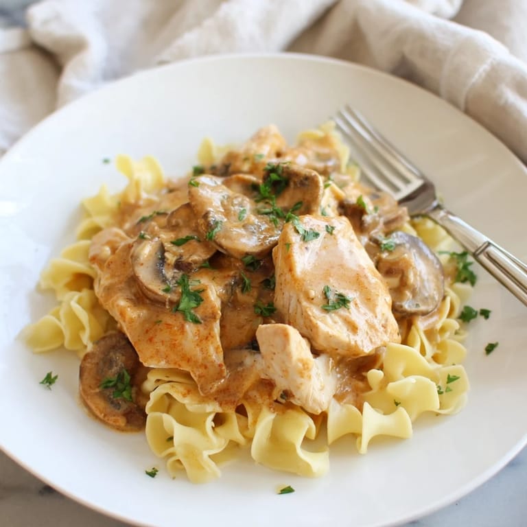 Comforting bowl of Chicken and Mushroom Stroganoff reveals golden chicken, browned mushrooms, and fresh parsley garnish, served alongside silky egg noodles on a rustic table.