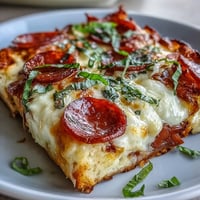 A close-up slice of High-Protein Pizza Breakfast Casserole reveals golden, bubbly mozzarella and cheddar over an egg layer, speckled with turkey pepperoni and diced red bell peppers. It's served on a white plate next to a small bowl of marinara for dipping, showcasing the hearty, meal-prep-friendly texture.